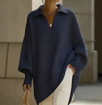 A person wearing an oversized navy blue knit polo-style sweater with wide draped sleeves, paired with white pants and layered gold necklaces, standing in a softly lit indoor setting.