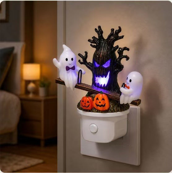 A Halloween-themed wall night light plugged into a wall outlet in a bedroom. The decorative plug-in light features a black gnarled tree with glowing purple eyes and mouth, two white ghosts wearing black bow ties and holding orange jack-o'-lanterns, and a brown branch structure.