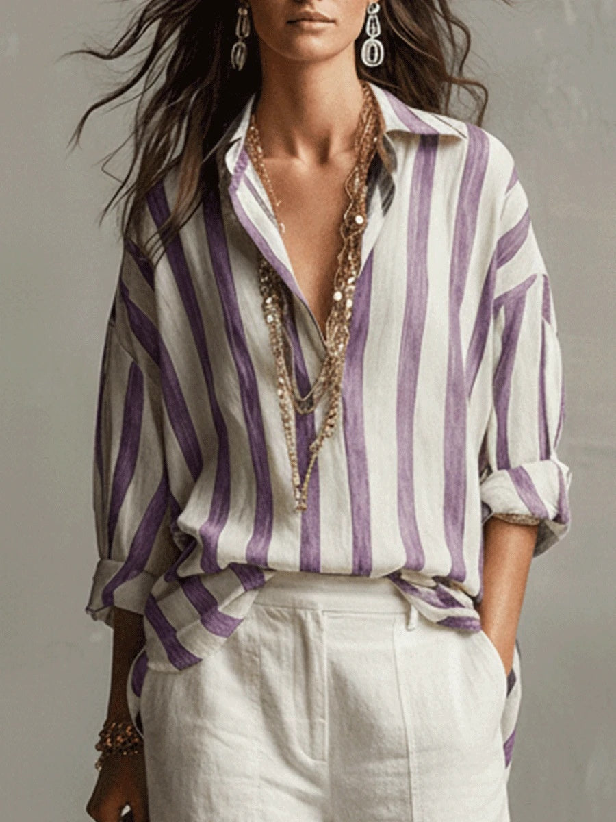 A woman wearing a loose white button-up shirt with vertical purple stripes, styled with layered gold necklaces and white trousers against a neutral gray background.