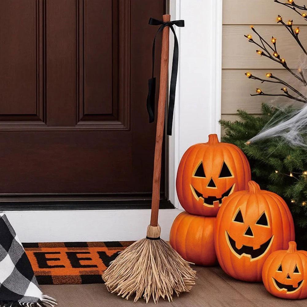 Halloween-themed front porch scene with a straw broom leaning against a dark brown door, three carved orange pumpkins, a small pumpkin, evergreen branches, cobwebs, and a plaid doormat.