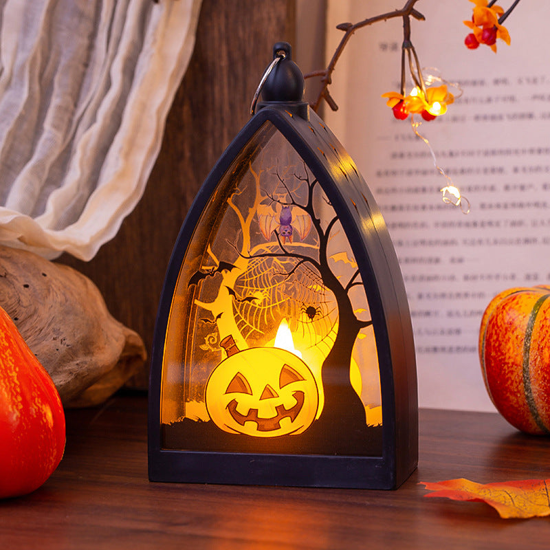 A Halloween-themed LED window box candle with a black frame and a glowing internal scene featuring a jack-o'-lantern, spiderweb, and bare tree branches, styled on a wooden surface with autumn props.