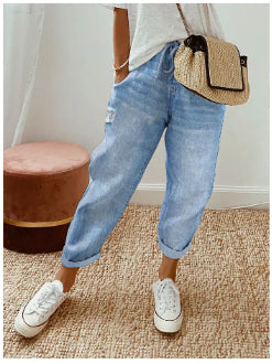 Person wearing blue jeans and white sneakers with a straw bag on a textured floor.