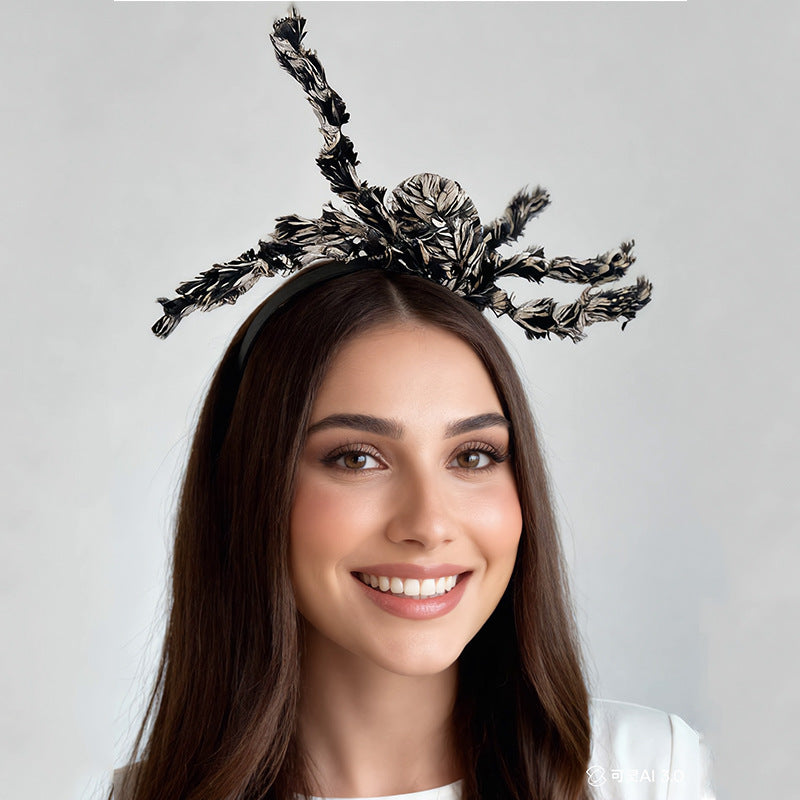 A smiling woman with long brown hair wears a large black and white feathered spider fascinator headpiece against a light gray studio background.