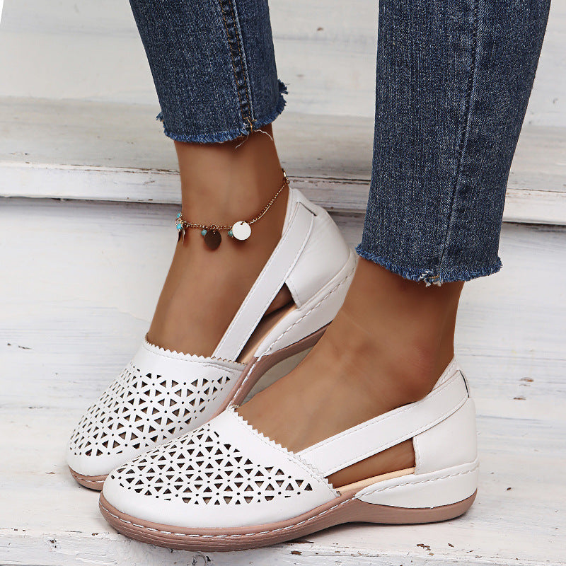 White sandals with cut-out design worn with blue jeans on a light wooden floor.