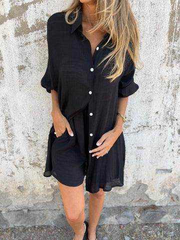 Woman wearing a black button-up shirt dress with pockets from ZA-Zola against a rustic wall