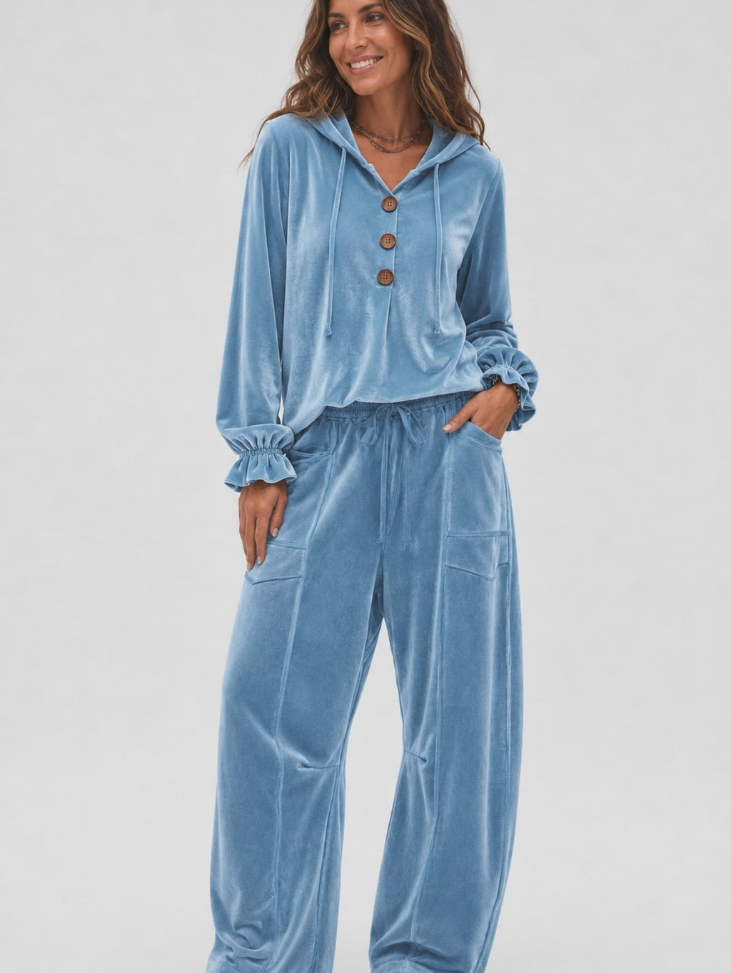 A woman modeling a light blue velvet two-piece loungewear set consisting of a long-sleeve button-front hooded top and matching wide-leg pants, against a plain light gray studio background.