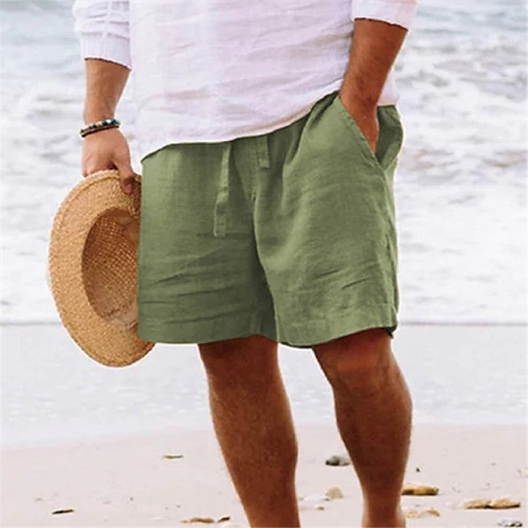 Person wearing green shorts and a white shirt on a beach, holding a straw hat.