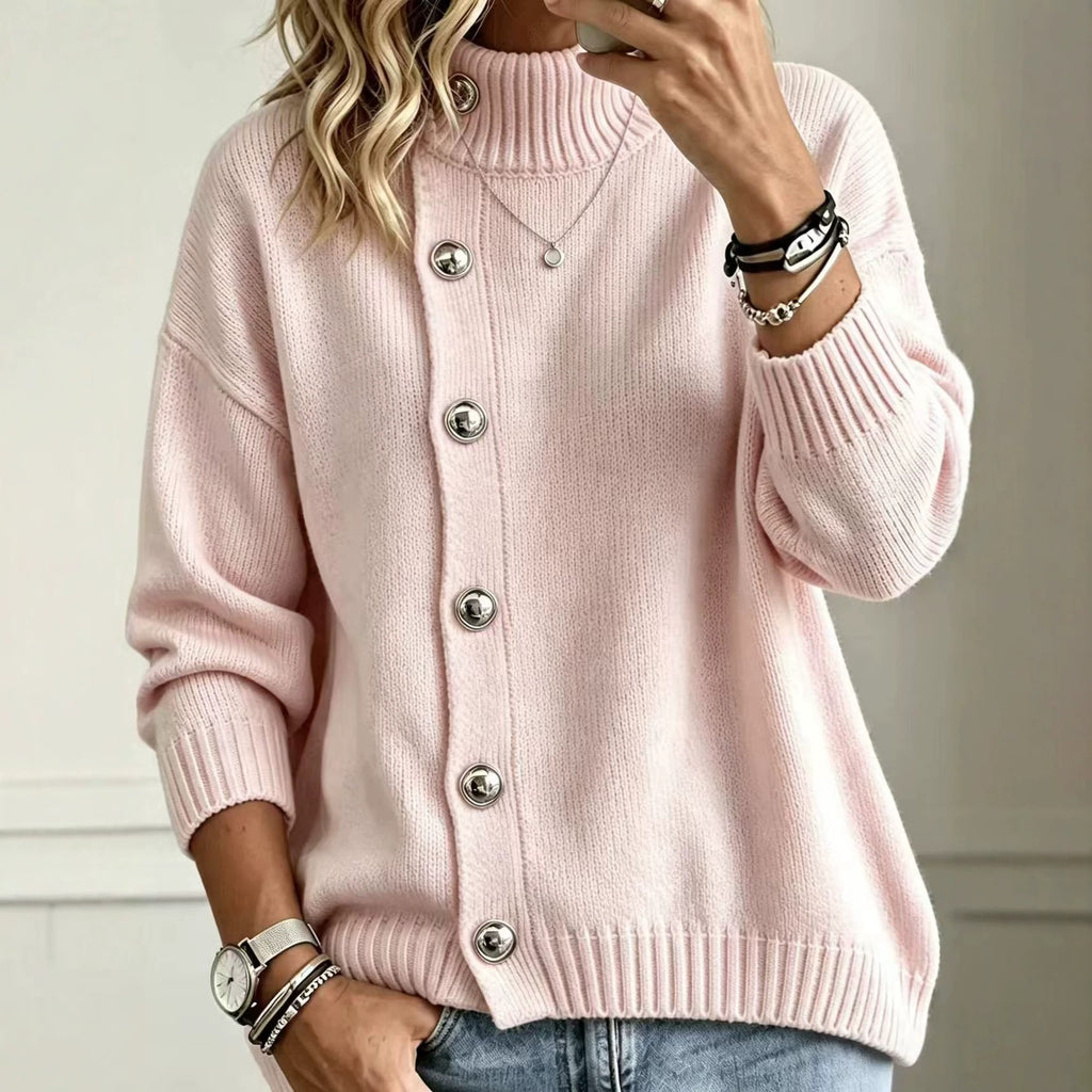 A woman wearing a light pink ribbed knit cardigan sweater with large silver buttons, paired with blue jeans and accessorized with bracelets and a necklace.
