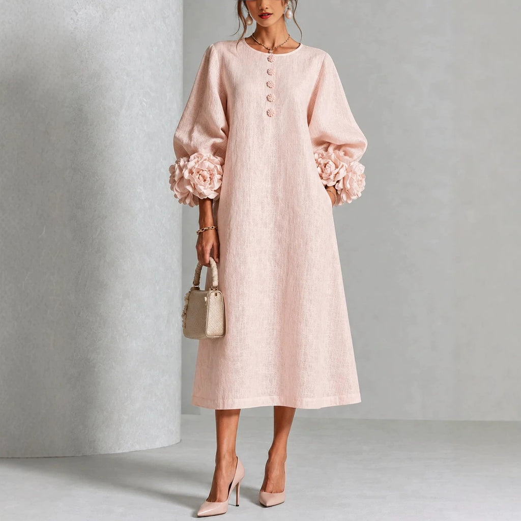 A woman models a light pink midi dress with voluminous long sleeves featuring large floral ruffle cuffs, a round neckline with decorative buttons, and a relaxed straight skirt. She is styled with a small beige handbag and beige high heels against a neutral gray studio backdrop.