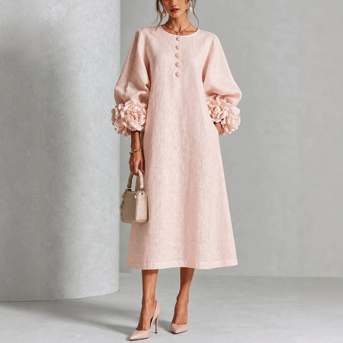 A woman models a light pink midi dress with voluminous long sleeves featuring large floral ruffle cuffs, a round neckline with decorative buttons, and a relaxed straight skirt. She is styled with a small beige handbag and beige high heels against a neutral gray studio backdrop.