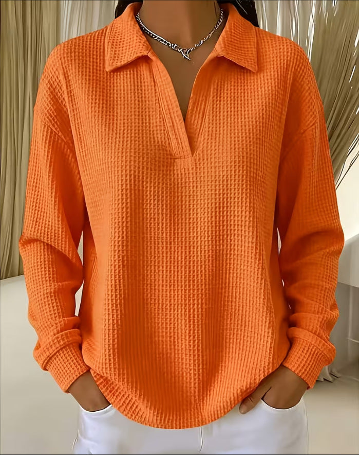A person wearing a bright orange waffle-knit long-sleeve collared top with a split V-neck, paired with white pants and a silver chain necklace.