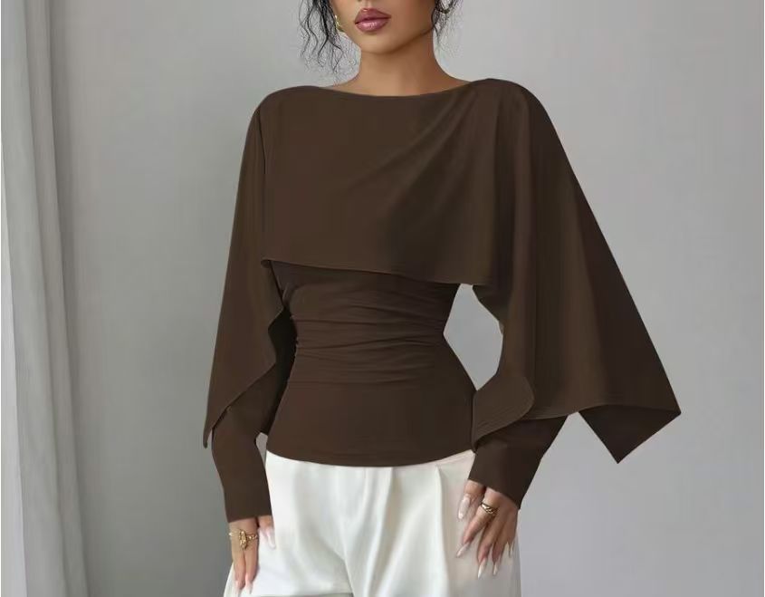 A woman wearing a dark brown long-sleeve top with a wide draped yoke and gathered waist, paired with white pants, against a plain gray background.