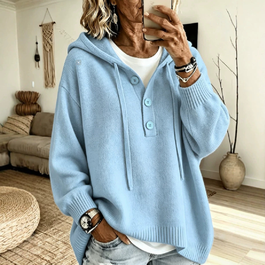 A woman takes a mirror selfie wearing a light blue knit hooded sweater with a buttoned henley-style front, layered over a white top and light denim jeans in a cozy living room setting.
