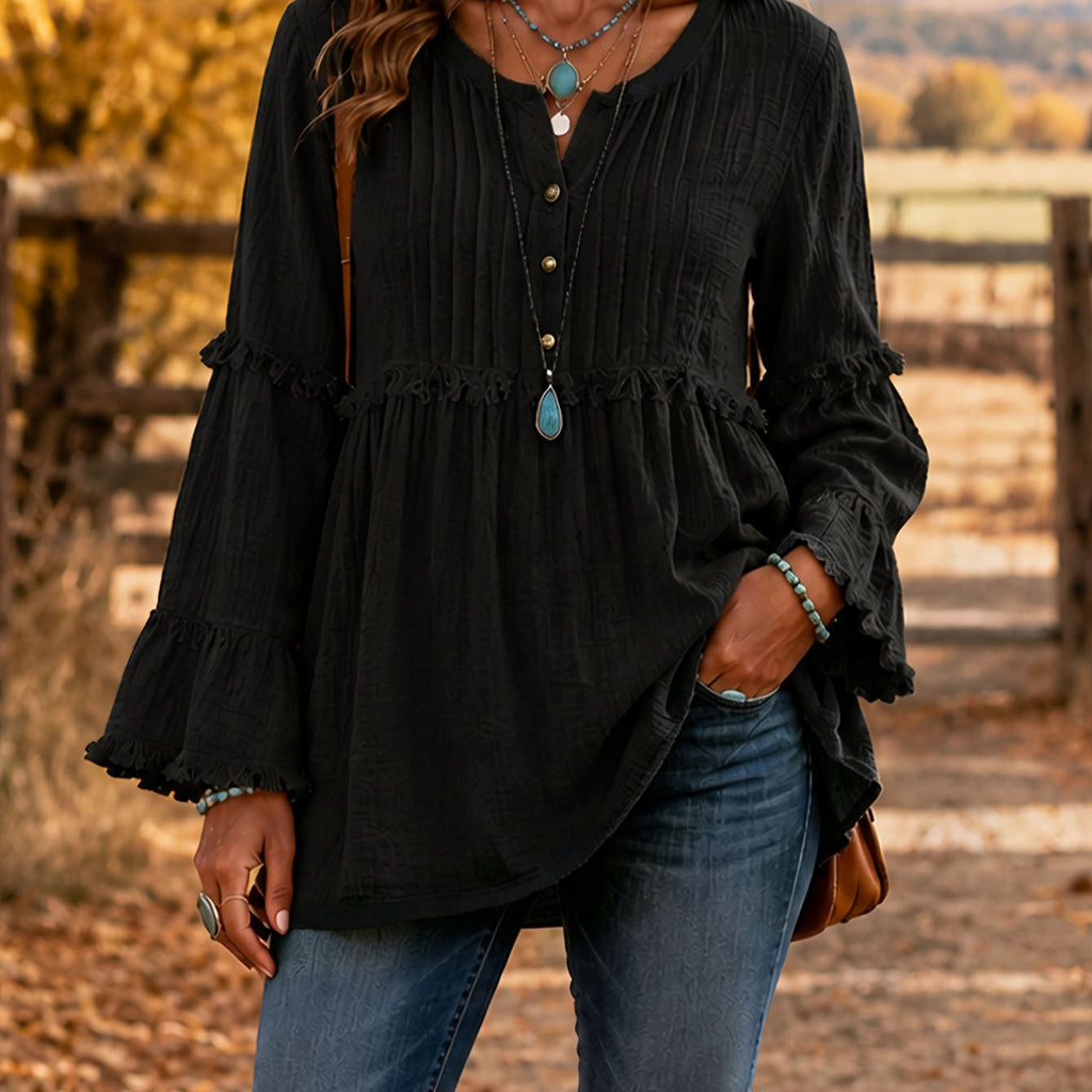 A woman wearing a black long-sleeve blouse with ruffled trim and a button placket, paired with blue jeans, standing outdoors in front of a rustic wooden fence.