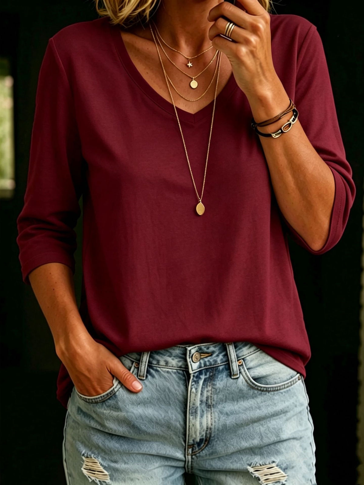 A woman wearing a burgundy long-sleeve V-neck top and light-wash distressed denim jeans, accessorized with layered gold necklaces and bracelets.