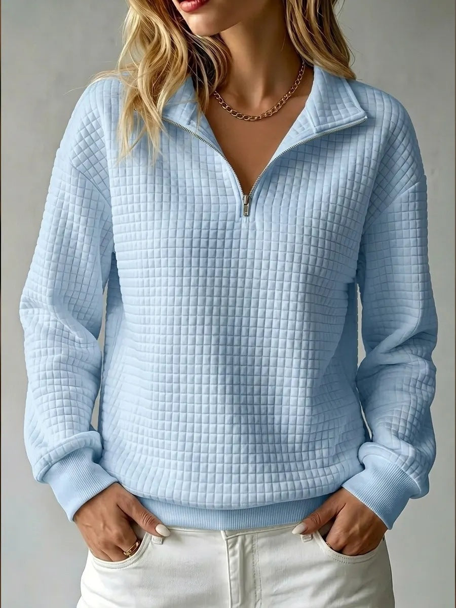 A woman wearing a light blue waffle-textured quarter-zip pullover sweatshirt with a stand collar, paired with white pants, shown against a neutral gray background.