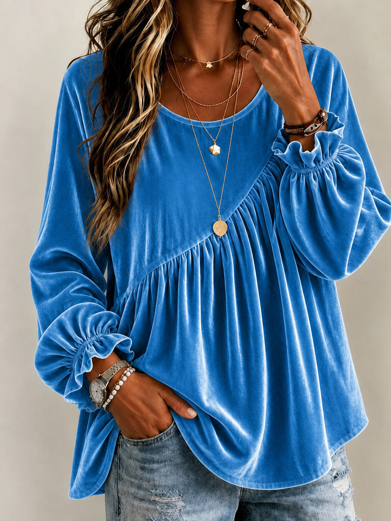 A woman wearing a bright blue velvet long-sleeve blouse with a gathered peplum-style waist and ruffled cuffs, paired with light-wash distressed jeans and layered gold necklaces.