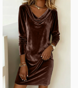 A woman wearing a long-sleeve cowl neck velvet dress in a deep chocolate brown color, accessorized with gold jewelry, standing against a neutral light gray wall.