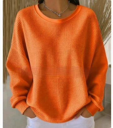 A person wearing a bright orange waffle-knit long-sleeve sweater with white pants, standing indoors against a neutral background.