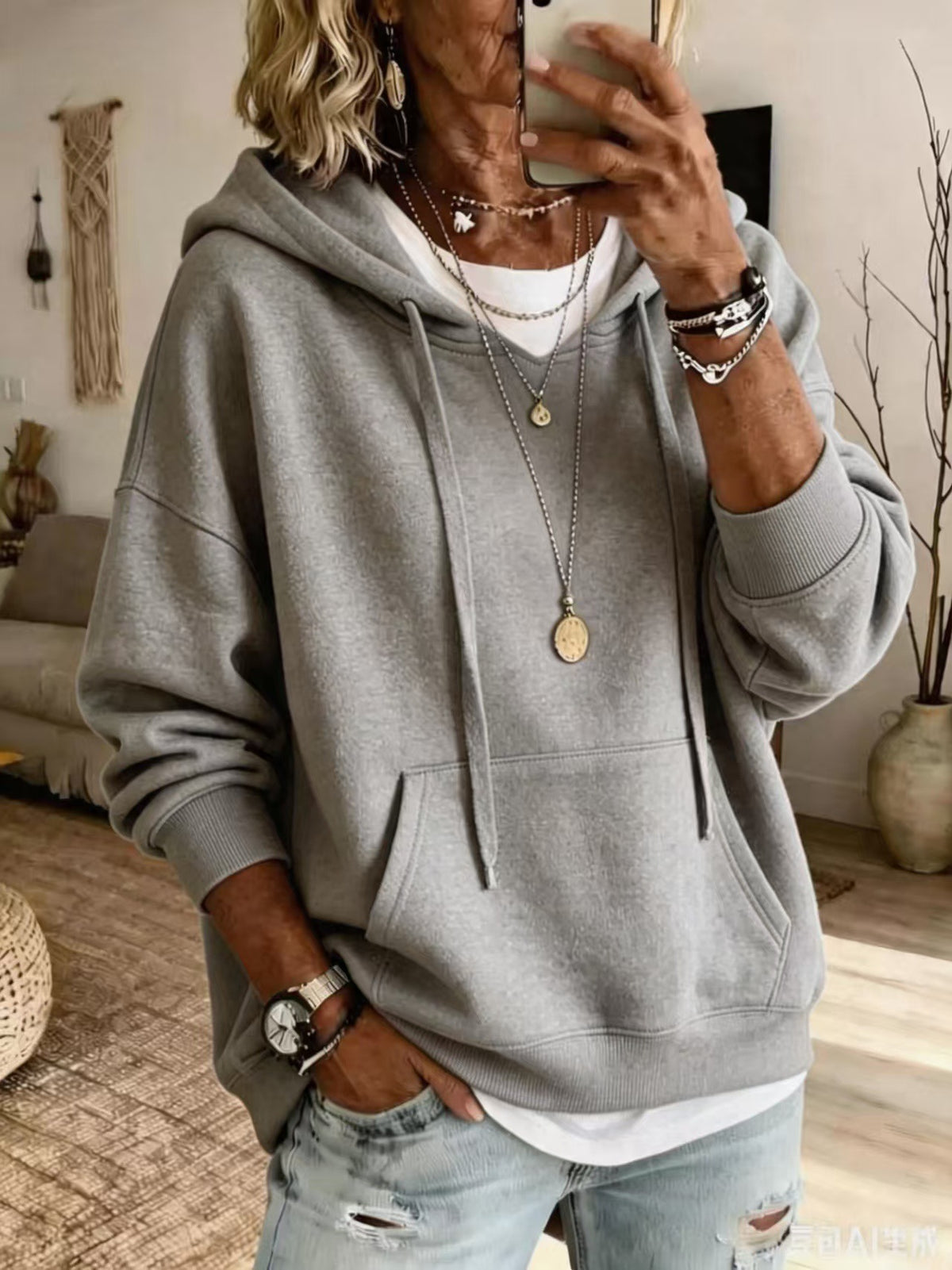 A woman takes a mirror selfie wearing a light gray oversized pullover hoodie with drawstrings, a white undershirt, and light-wash distressed jeans. She is accessorized with layered necklaces, bracelets, and a watch.