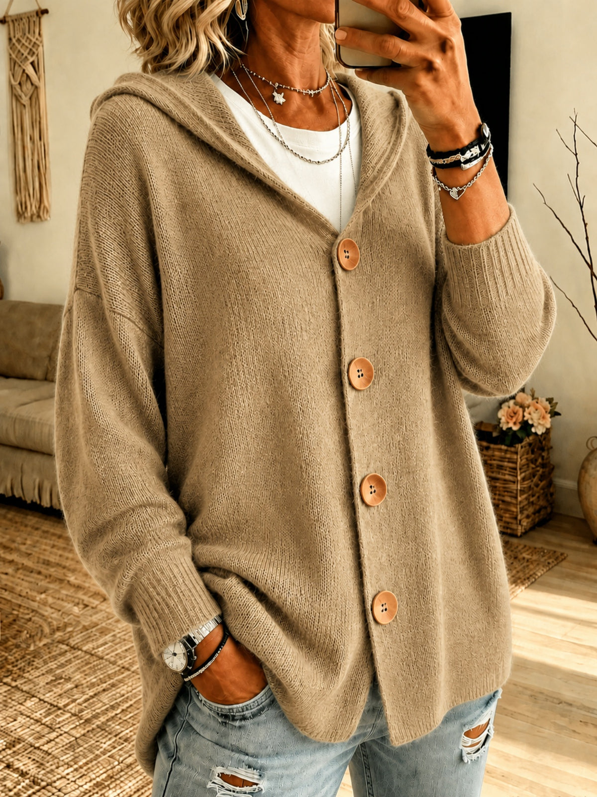 A woman wearing a beige oversized knit cardigan with a hood and large brown buttons, layered over a white top and paired with distressed light blue jeans in a cozy indoor setting.