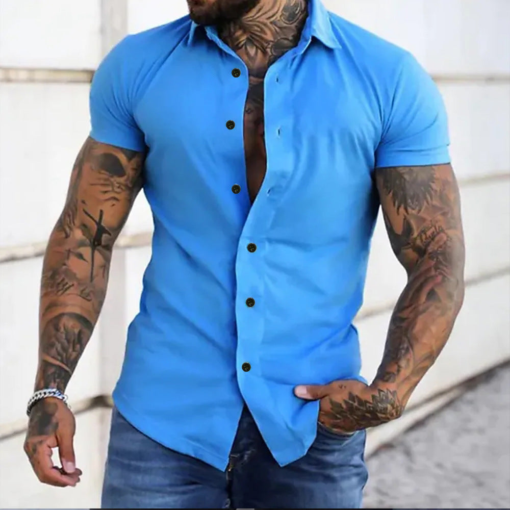 Man wearing a blue short-sleeve button-up shirt with tattoos on arms, standing against a light background.