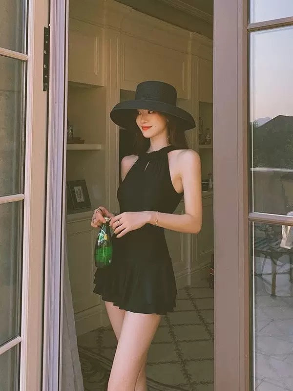 Woman in a black dress and hat holding a green bottle, standing in a doorway.