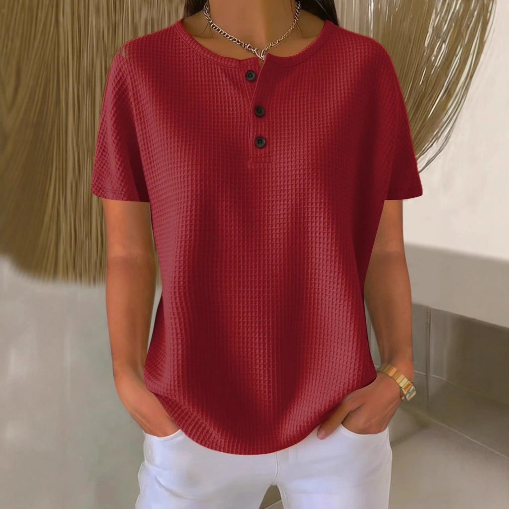 Person wearing a red textured shirt with black buttons and white pants in a neutral setting