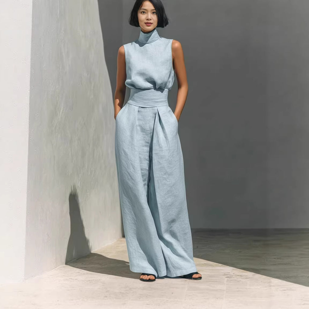 Woman wearing a light blue sleeveless top and wide-leg pants against a gray wall.