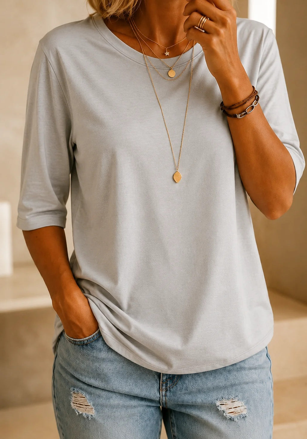 A woman wearing a light gray heathered short-sleeve t-shirt with rolled-up sleeves and light-wash distressed denim jeans, accessorized with layered gold necklaces and bracelets.