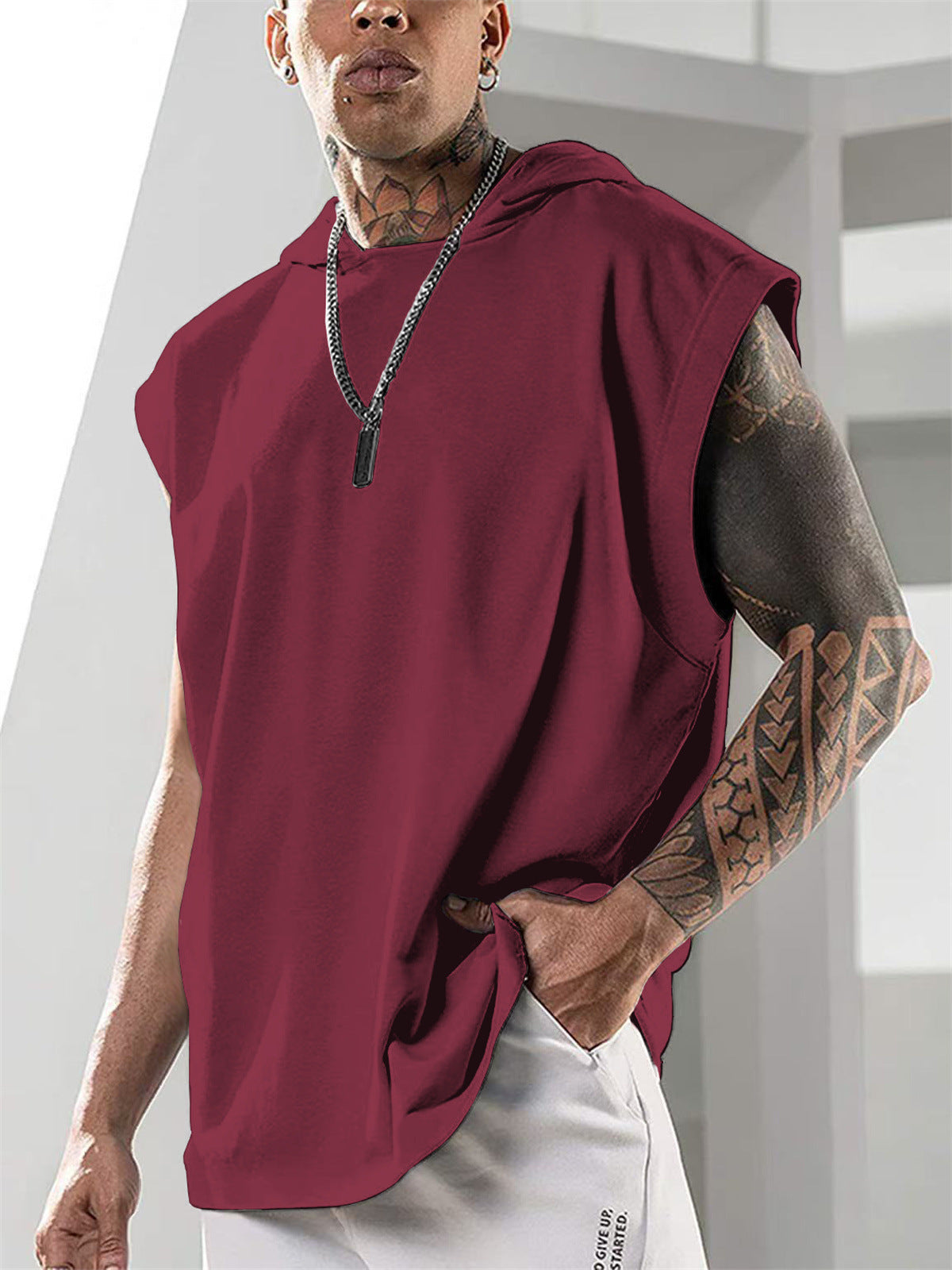 Person wearing a maroon sleeveless shirt with tattoos on arms, standing against a light background.