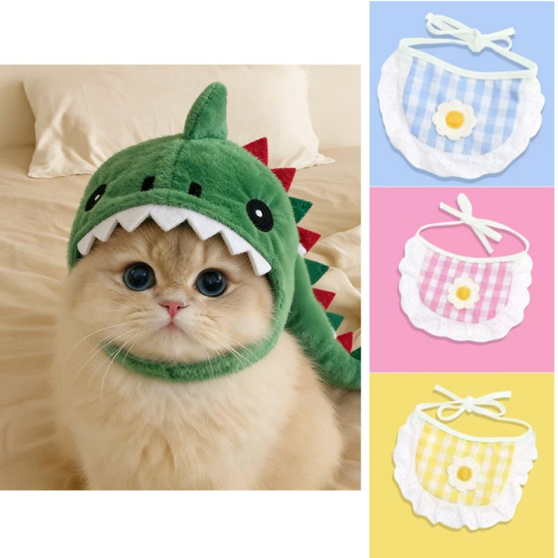 A fluffy cream-colored cat wearing a green dinosaur costume hat with white teeth and red spikes, shown on a bed. The right side shows three color variants of a matching bib: blue-and-white gingham with a yellow flower, pink-and-white gingham with a yellow flower, and yellow-and-white gingham with a yellow flower.