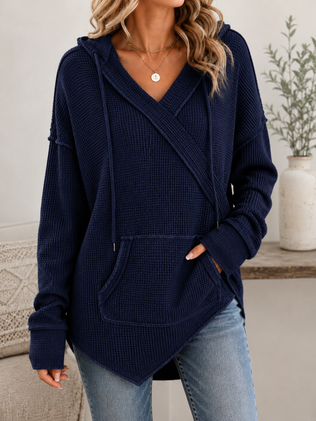 A woman wearing a navy blue waffle-knit hooded sweater with a wrap-style front, drawstrings, and front pockets, paired with light blue jeans in a softly styled indoor setting.