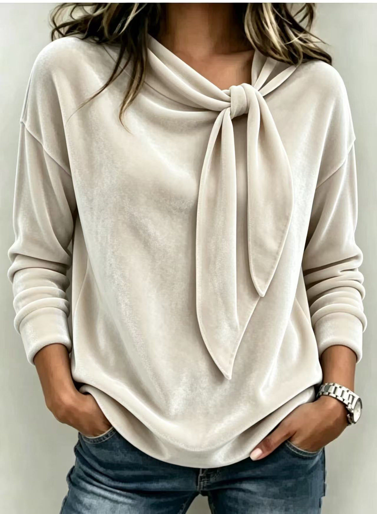 A woman wearing a light beige long-sleeve knit top with a large draped tie detail at the neckline, paired with blue jeans.