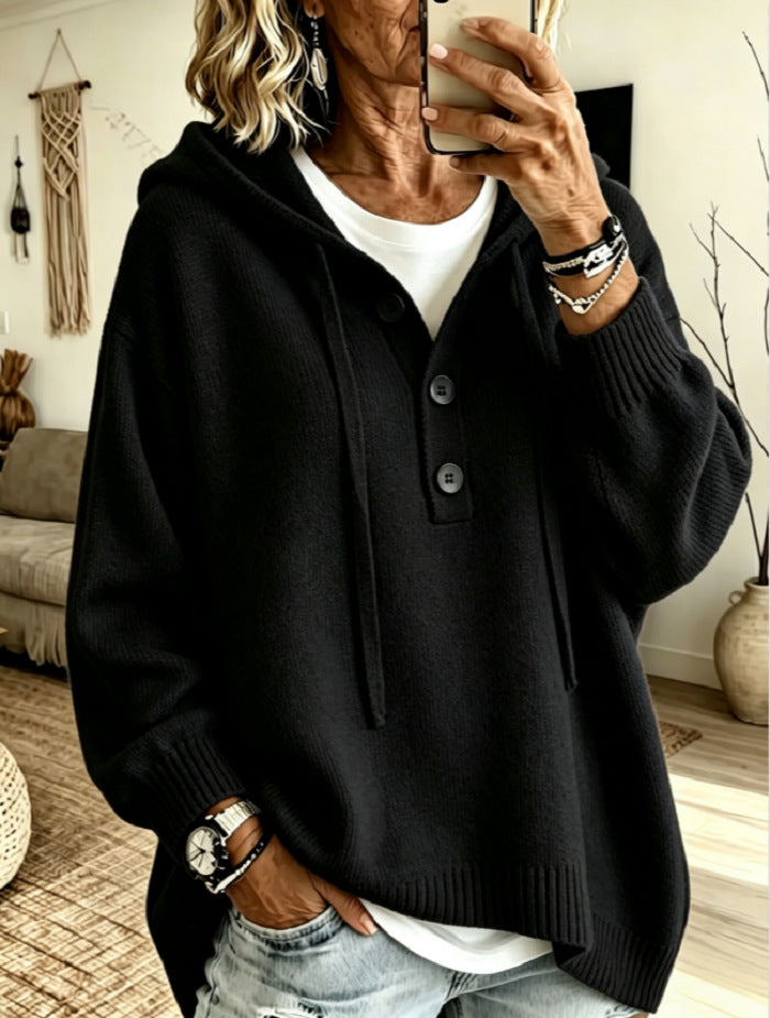 A woman takes a mirror selfie wearing a black knit hooded sweater with a buttoned placket over a white t-shirt and light-wash jeans, in a cozy living room setting.