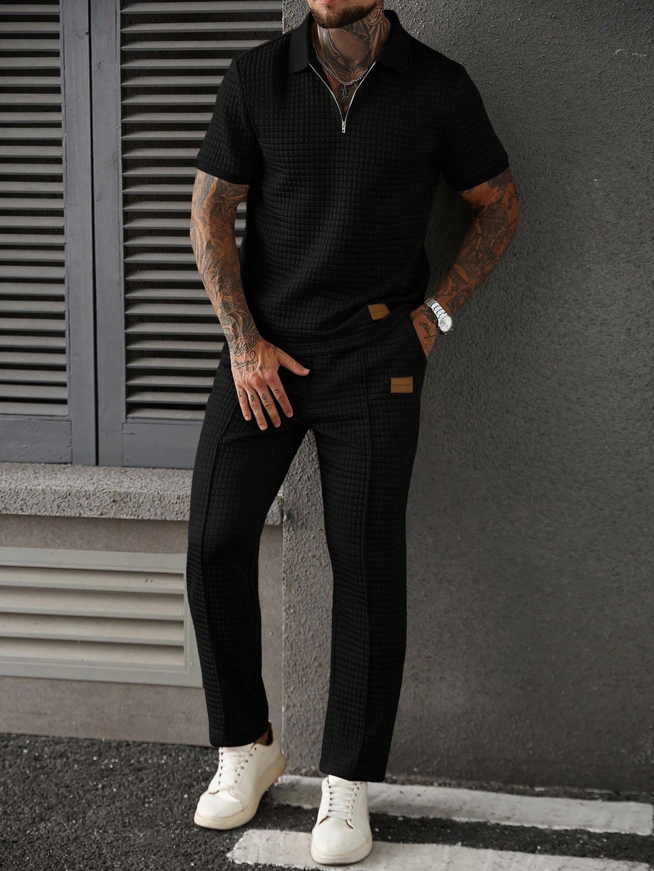 Man wearing a black polo shirt and pants with white sneakers against a gray wall.