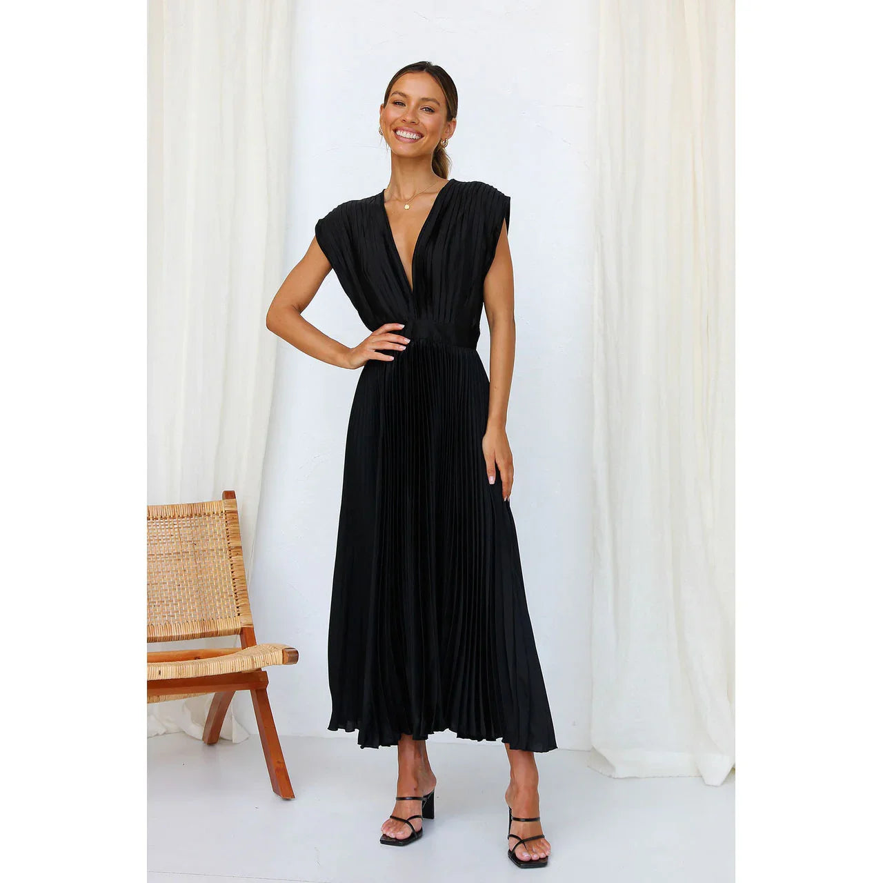 Woman wearing a black dress standing against a white curtain with a wooden chair.
