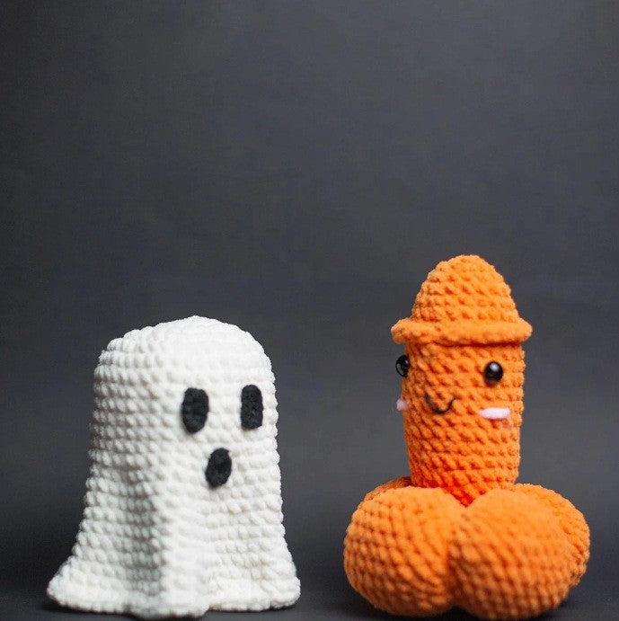 Two handmade crocheted plush toys on a dark gray background: a white ghost with black eyes and mouth, and an orange phallus-shaped figure wearing an orange hat with a smiling face.