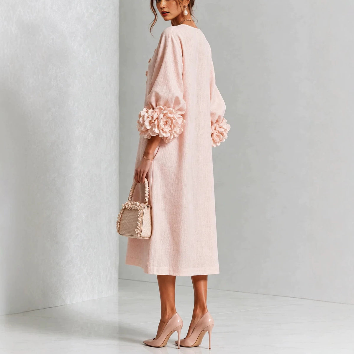 A woman models a light pink midi dress with voluminous sleeves and large floral embellishments at the cuffs, paired with nude heels and a matching pink handbag against a minimalist gray studio background.