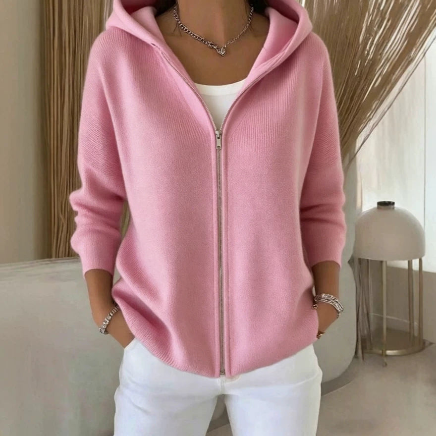 A woman wearing a light pink zip-up hoodie with a ribbed texture, layered over a white top and white pants, standing in a softly lit indoor setting.