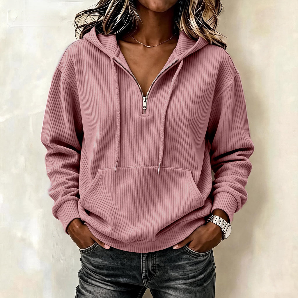A woman wearing a ribbed pink half-zip hoodie with a drawstring hood and kangaroo pocket, paired with dark gray jeans, standing against a light beige textured wall.