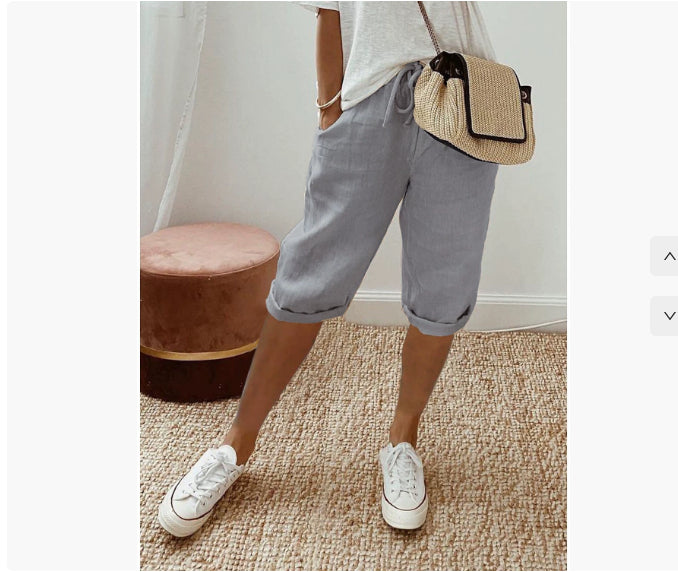 Person wearing gray shorts, white sneakers, and a straw bag in a room with a pink ottoman.