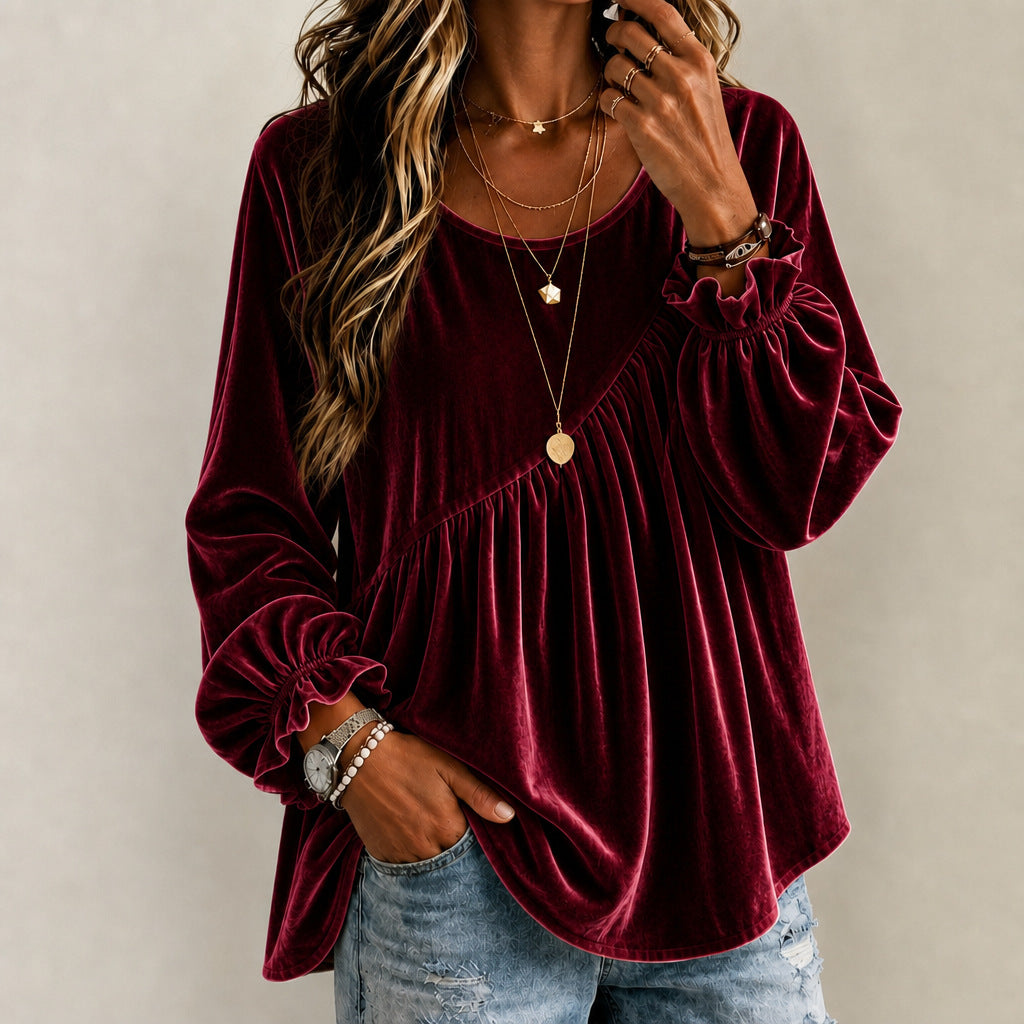 A woman wearing a long-sleeve burgundy velvet top with a gathered peplum-style waist and elasticized cuffs, paired with light-wash distressed jeans and layered gold necklaces, against a neutral beige background.