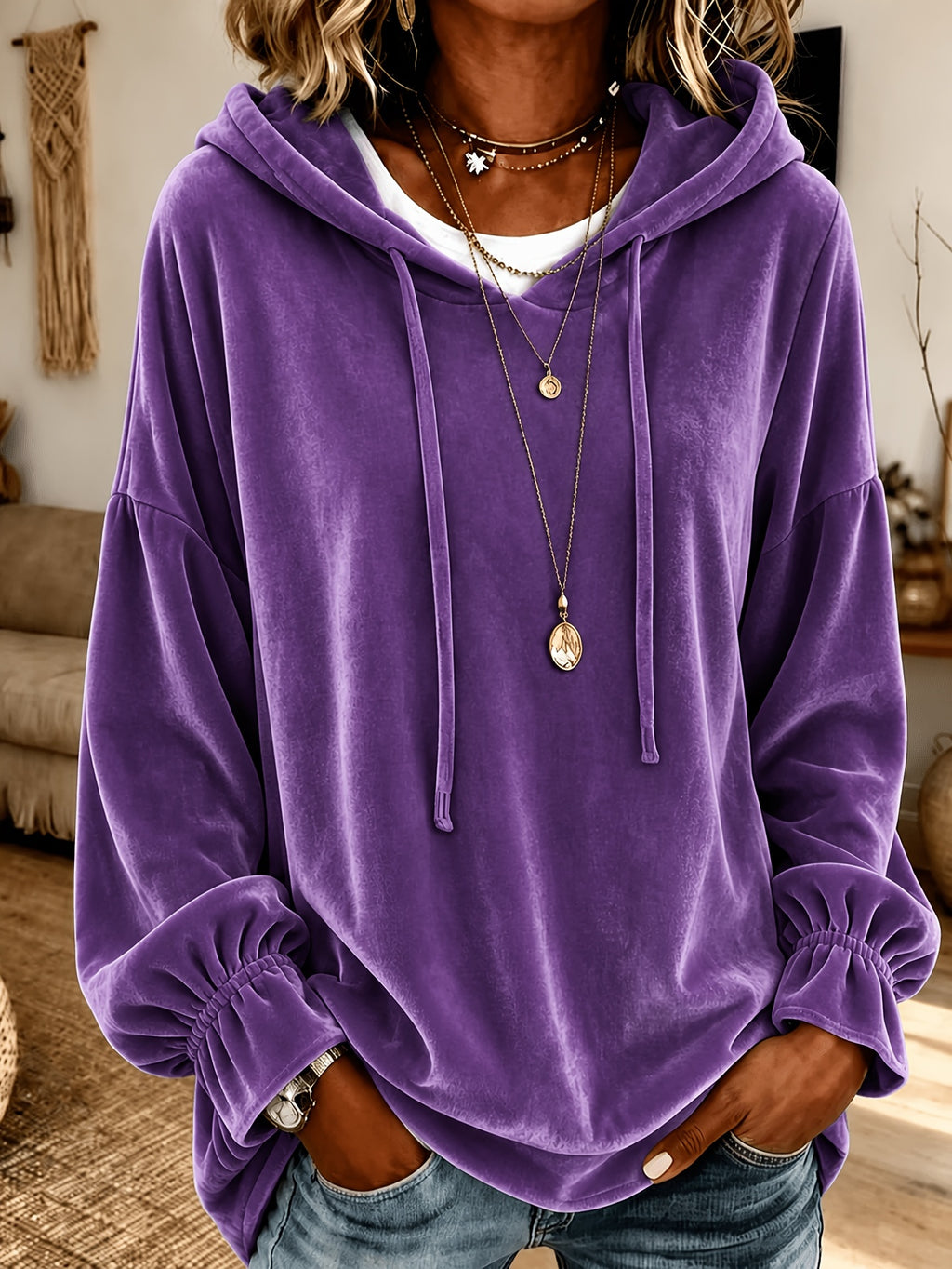 A person wearing an oversized purple velvet hoodie with drawstrings and elasticized cuffs, layered over a white shirt and accessorized with layered necklaces, in a cozy indoor setting.
