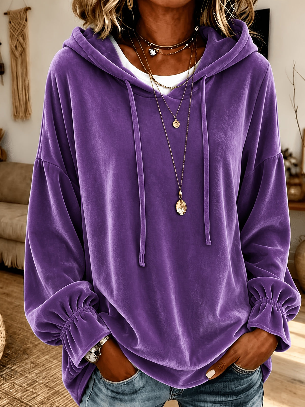 A person wearing an oversized purple velvet hoodie with drawstrings and elasticized cuffs, layered over a white shirt and accessorized with layered necklaces, in a cozy indoor setting.