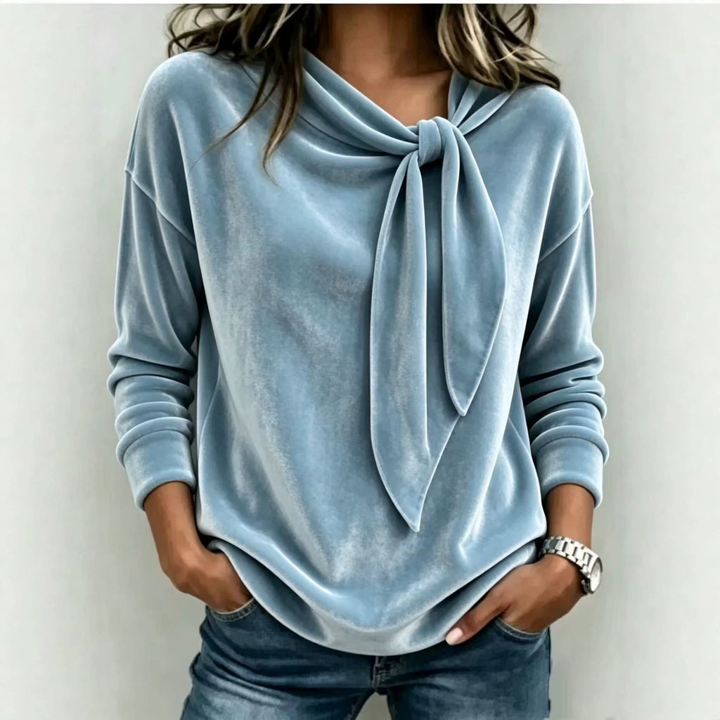 A woman wearing a light blue velvet long-sleeve top with a knotted tie neckline, paired with blue jeans, shown against a plain light gray background.