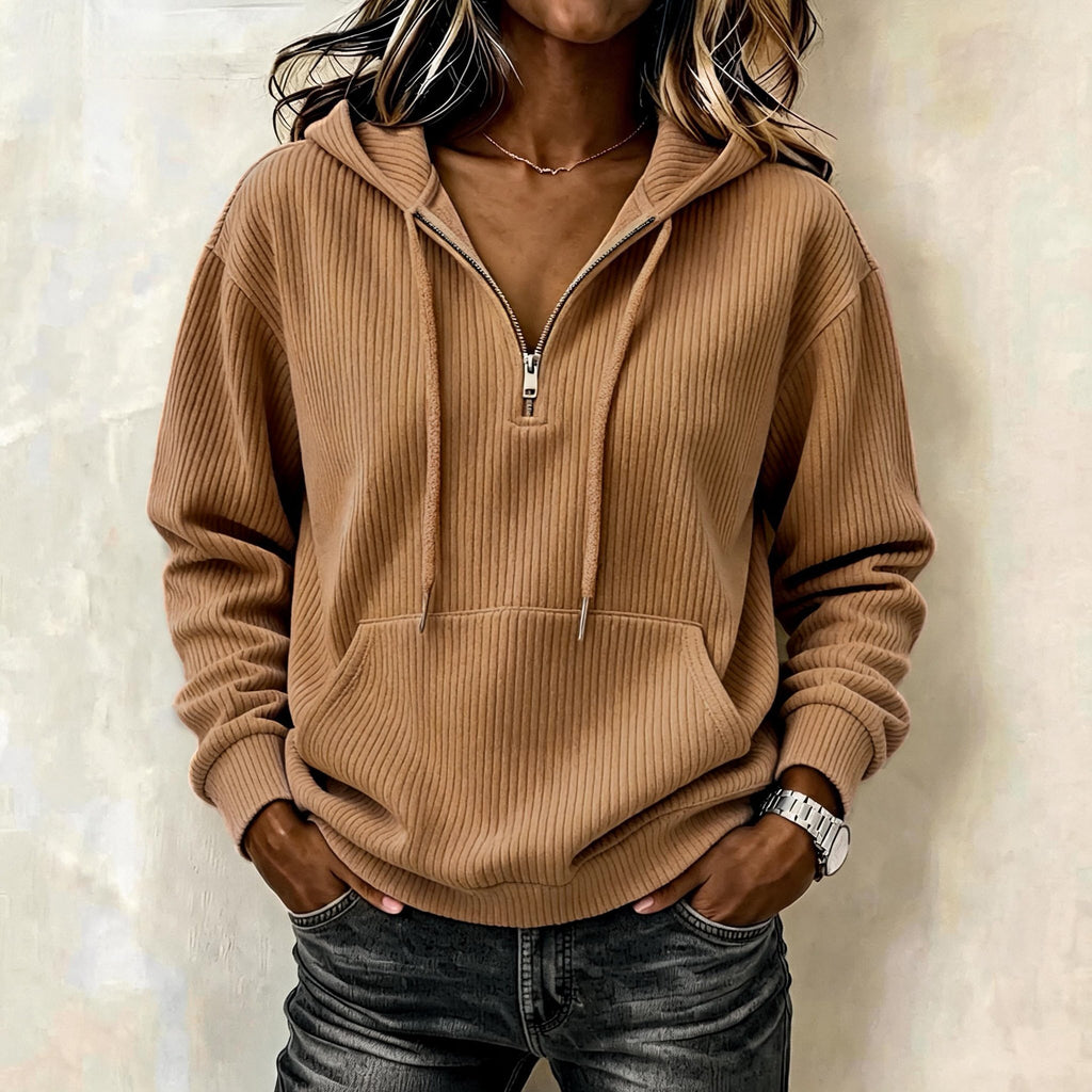 A woman wearing a ribbed brown half-zip hoodie with a drawstring hood and kangaroo pocket, paired with dark gray jeans, standing against a textured beige wall.