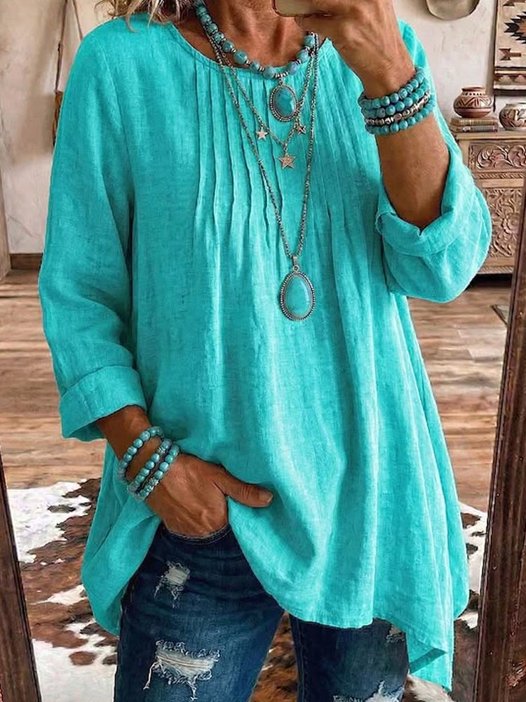 A woman takes a mirror selfie wearing a loose-fitting, long-sleeve turquoise linen top with a gathered neckline, paired with distressed blue jeans and layered silver jewelry.