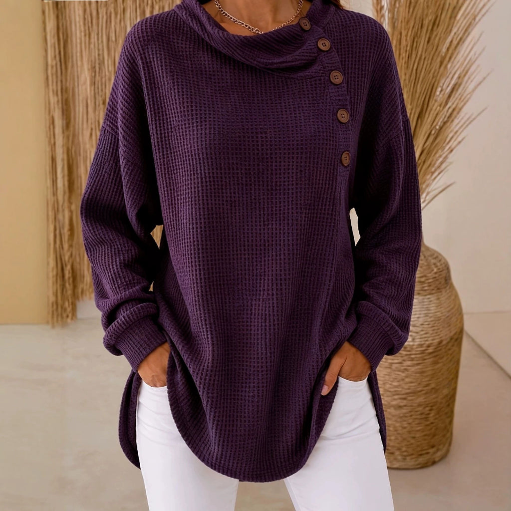 A woman wearing a dark purple waffle-knit cowl-neck sweater with brown buttons on the side, paired with white pants, standing in a neutral indoor setting.