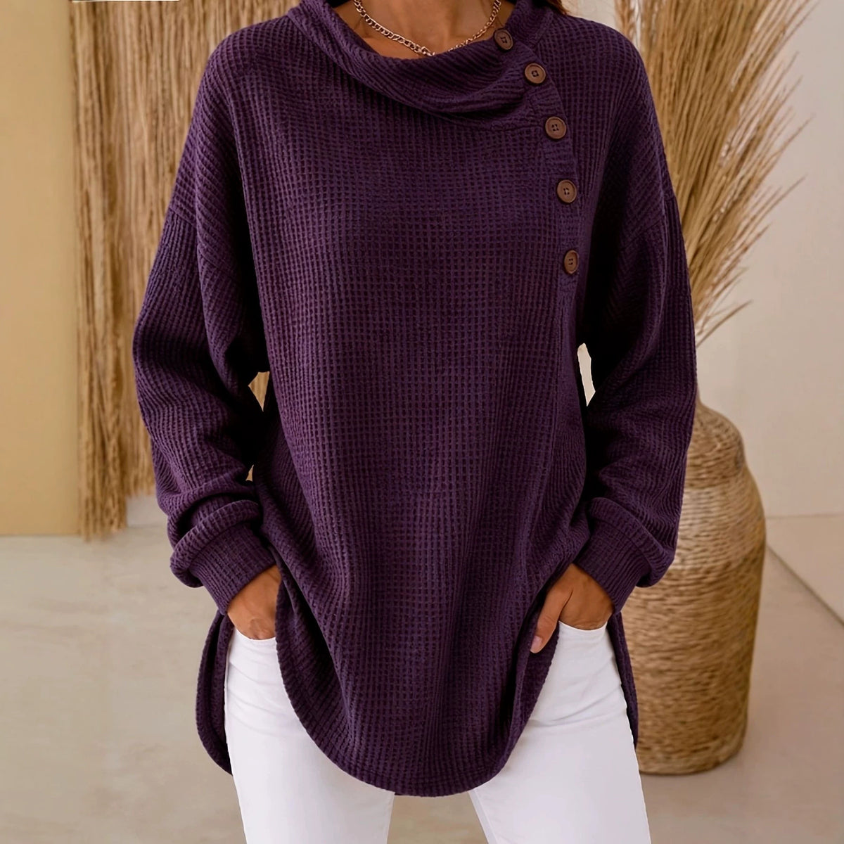 A woman wearing a dark purple waffle-knit cowl-neck sweater with brown buttons on the side, paired with white pants, standing in a neutral indoor setting.